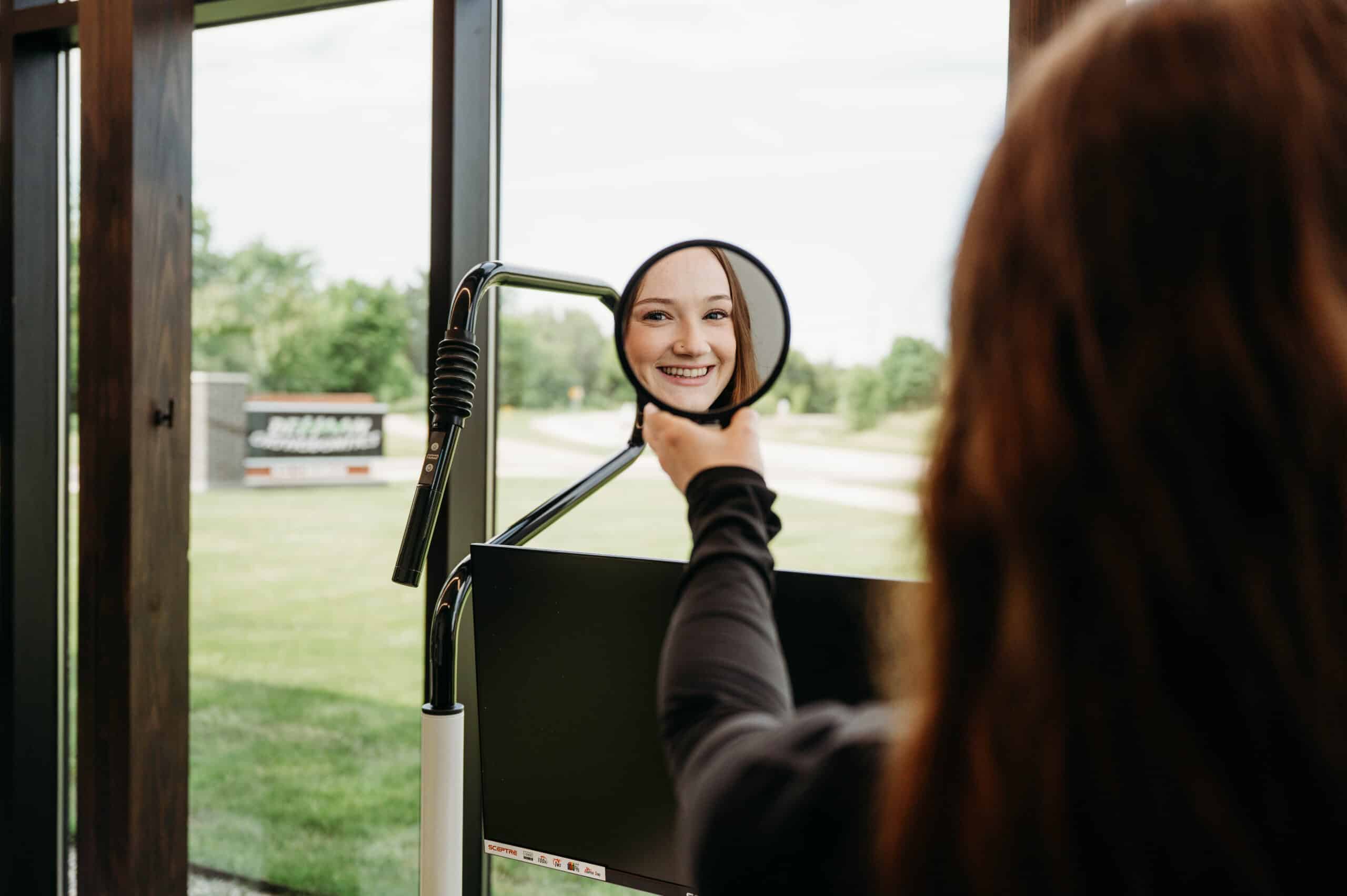 Patient smiling at her reflection in a handheld mirror after treatment in the dental chair at DeHaan Orthodontics in Lake Orion, MI