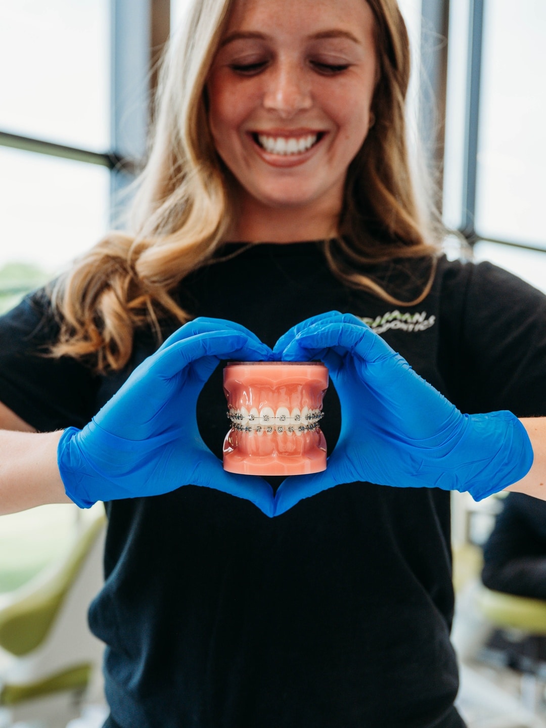 Team member smiling and forming a heart shape with her gloved hands around a dental model with braces at DeHaan Orthodontics in Lake Orion, MI