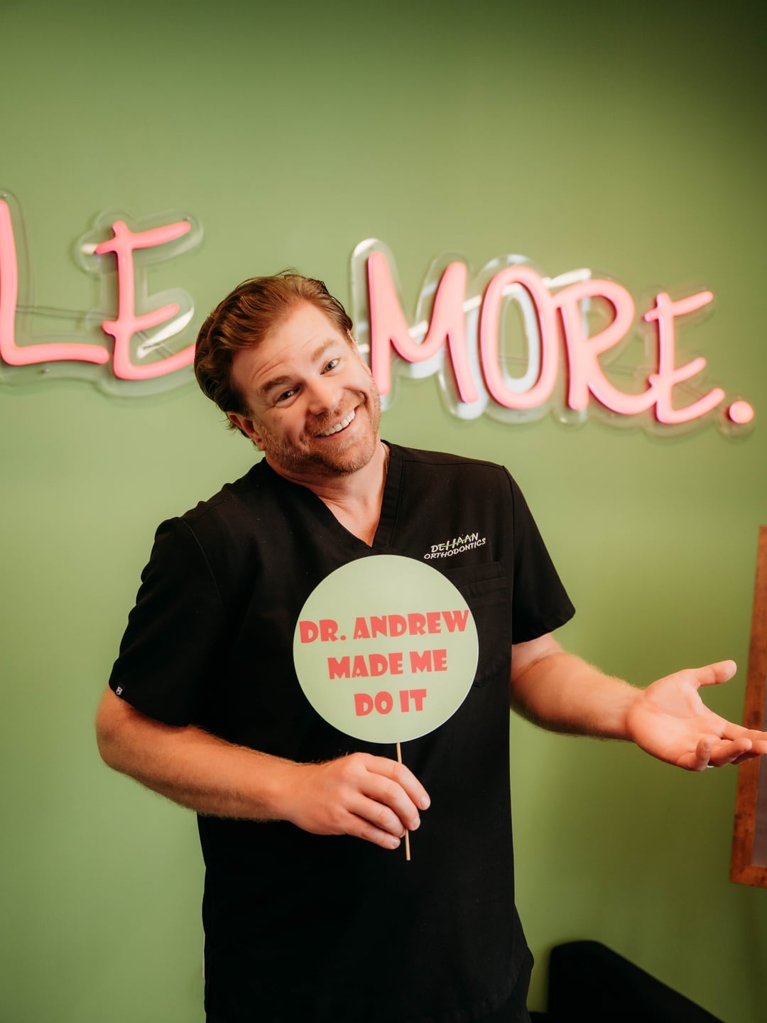 Dr. Andrew DeHaan smiling and holding a sign that reads Dr. Andrew Made Me Do It in front of the Smile More wall at DeHaan Orthodontics in Lake Orion, MI