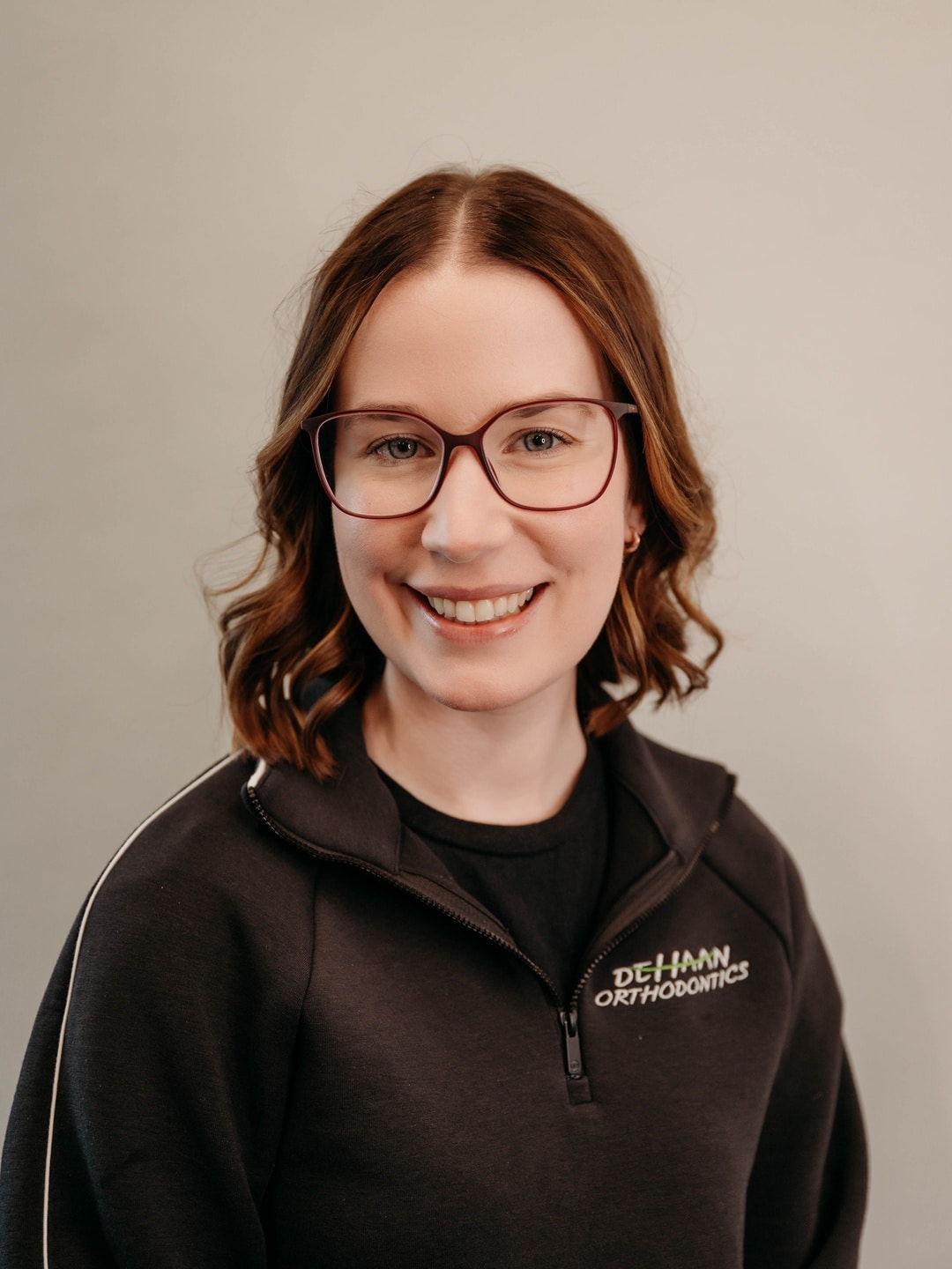Sam, Insurance Coordinator at DeHaan Orthodontics, smiling in a branded team jacket at DeHaan Orthodontics in Lake Orion, MI