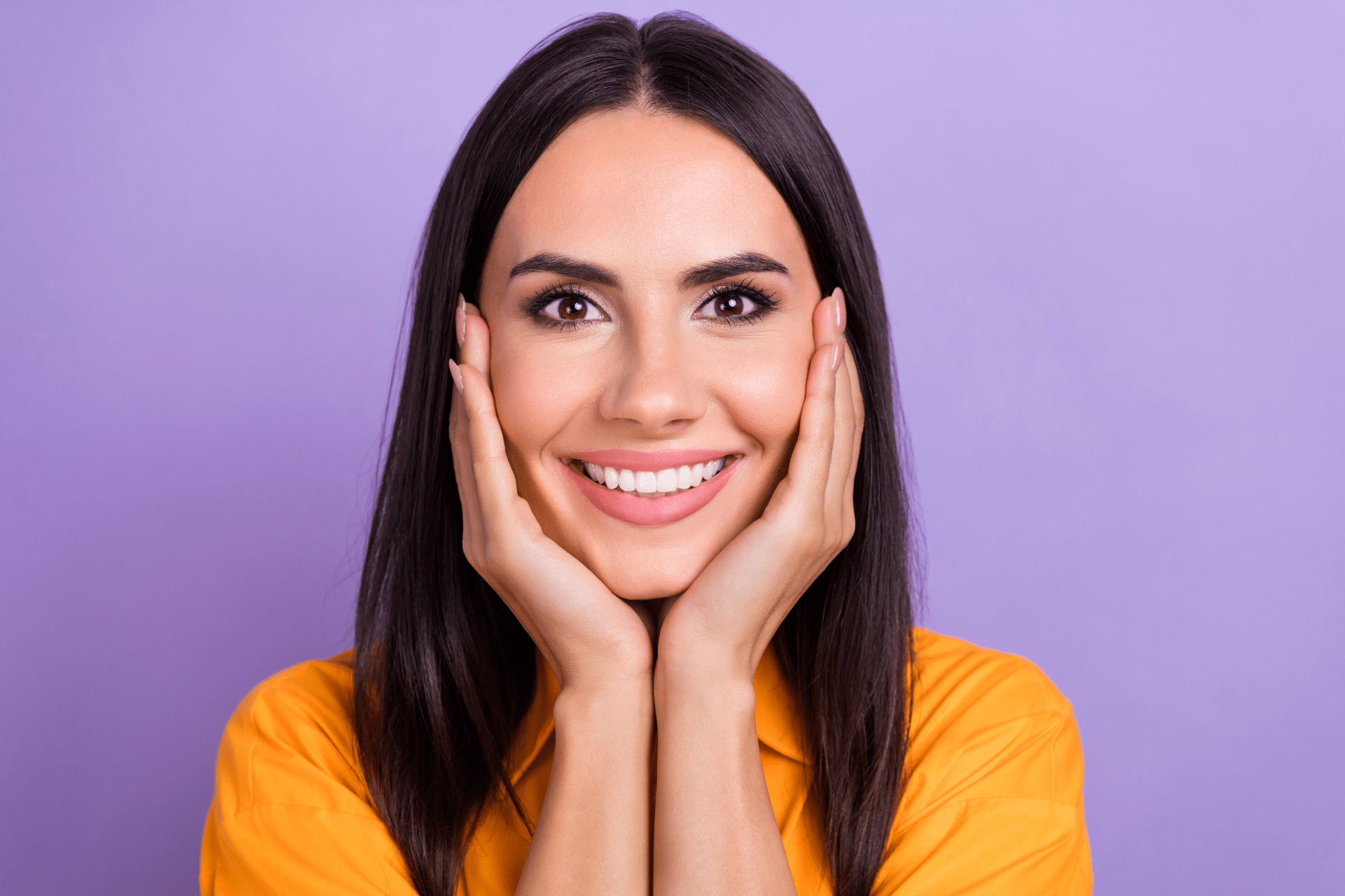 Smiling woman resting both hands on her cheeks against a purple background.